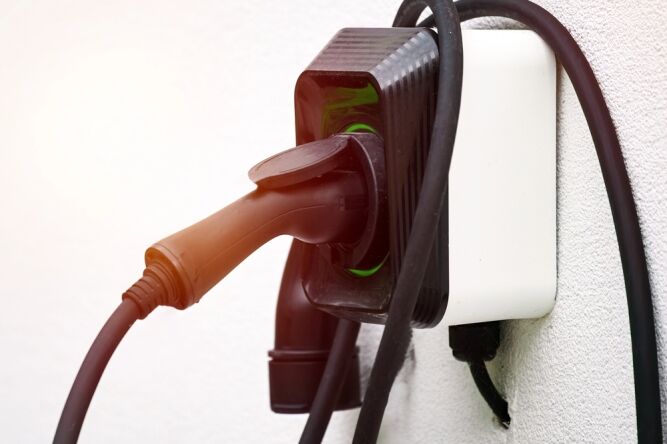 Close-up of an electric vehicle charging plug connected to a black charging station mounted on a white wall, with cables extending from the device.