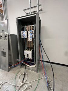 An open electrical panel with exposed circuit breakers and multiple colored cables connected, situated against a plain wall on a concrete floor.