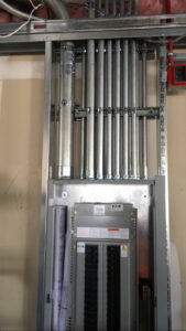 Electrical panel with multiple metal conduit pipes above, circuit breakers visible on the panel, wiring diagrams to the side, and a vertical measuring stick on the right.