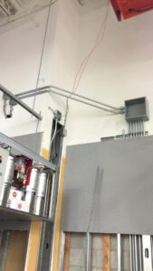 Exposed electrical conduit and cables run along a white wall and ceiling corner; metal pipes, fire suppression cylinders, and unfinished drywall are visible.