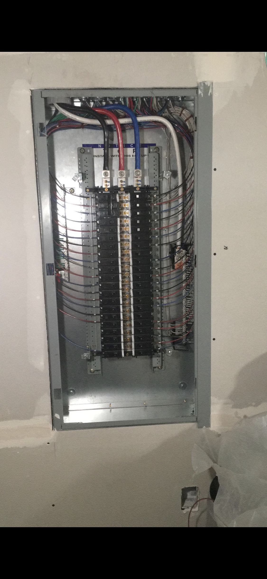 Open electrical panel with multiple circuit breakers and colored wires installed on a wall with unfinished drywall surrounding the panel.