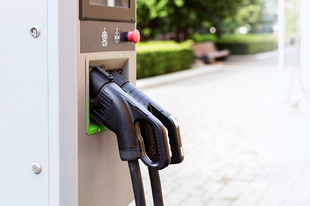 Close-up of an electric vehicle charging station with two charging connectors plugged into the unit; a park bench and greenery are visible in the background.