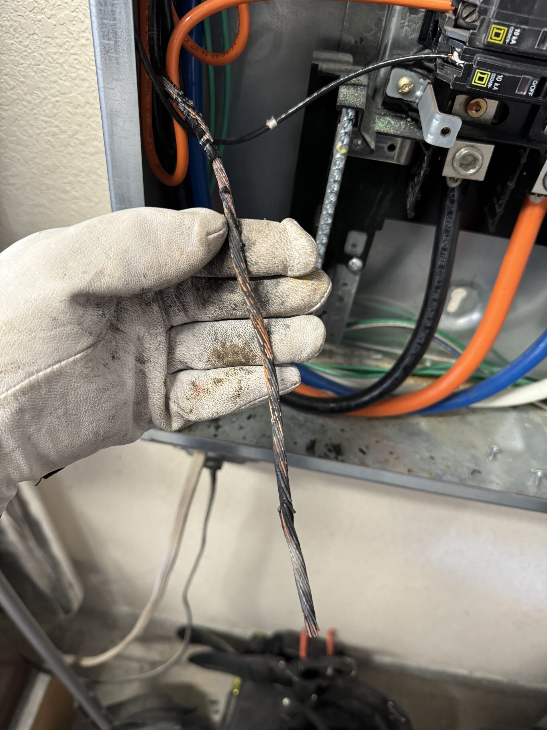 A gloved hand holds a damaged, partially melted electrical wire inside a circuit breaker panel with visible wiring.