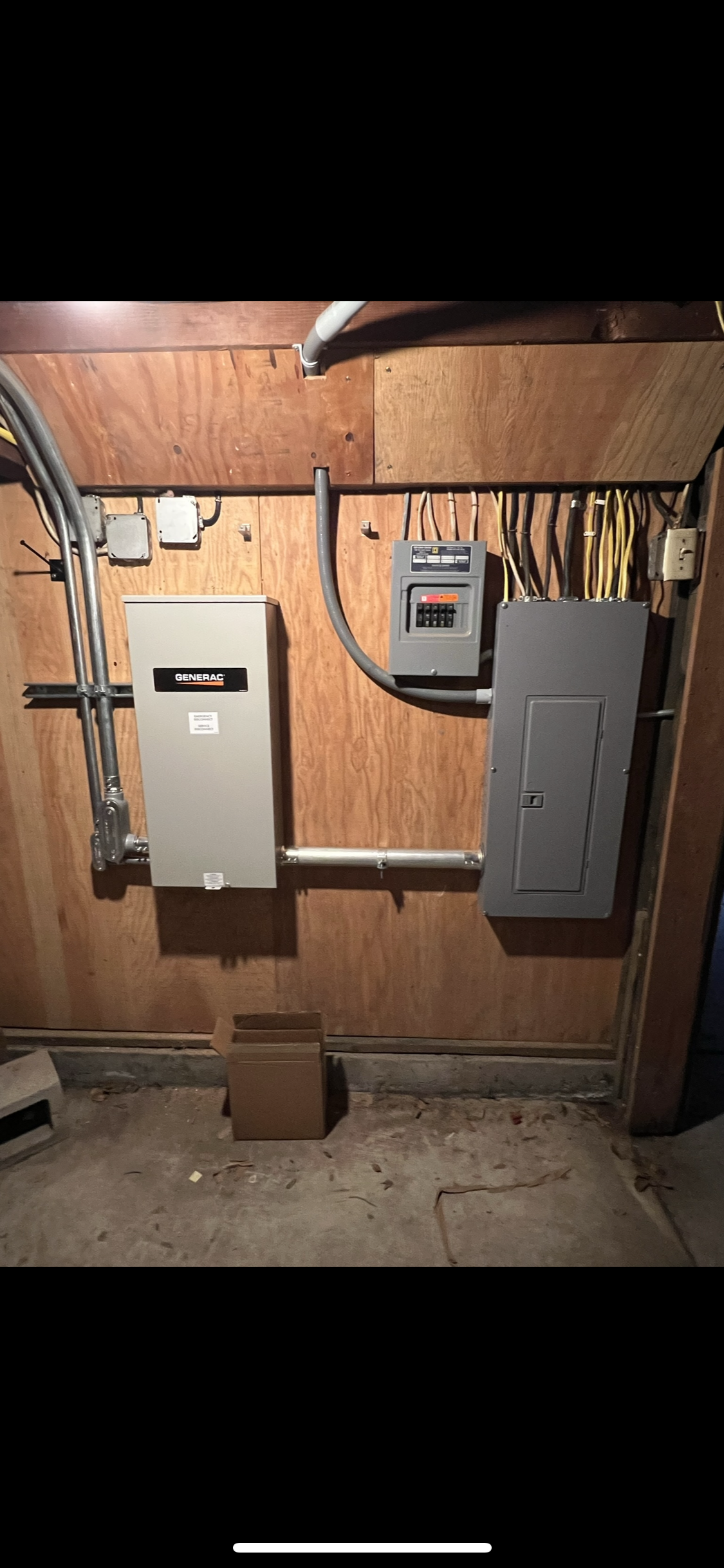 Electrical panel setup on a wooden wall, featuring boxes labeled Generac and another electrical panel with visible wiring and conduit. An open cardboard box sits on the floor below.