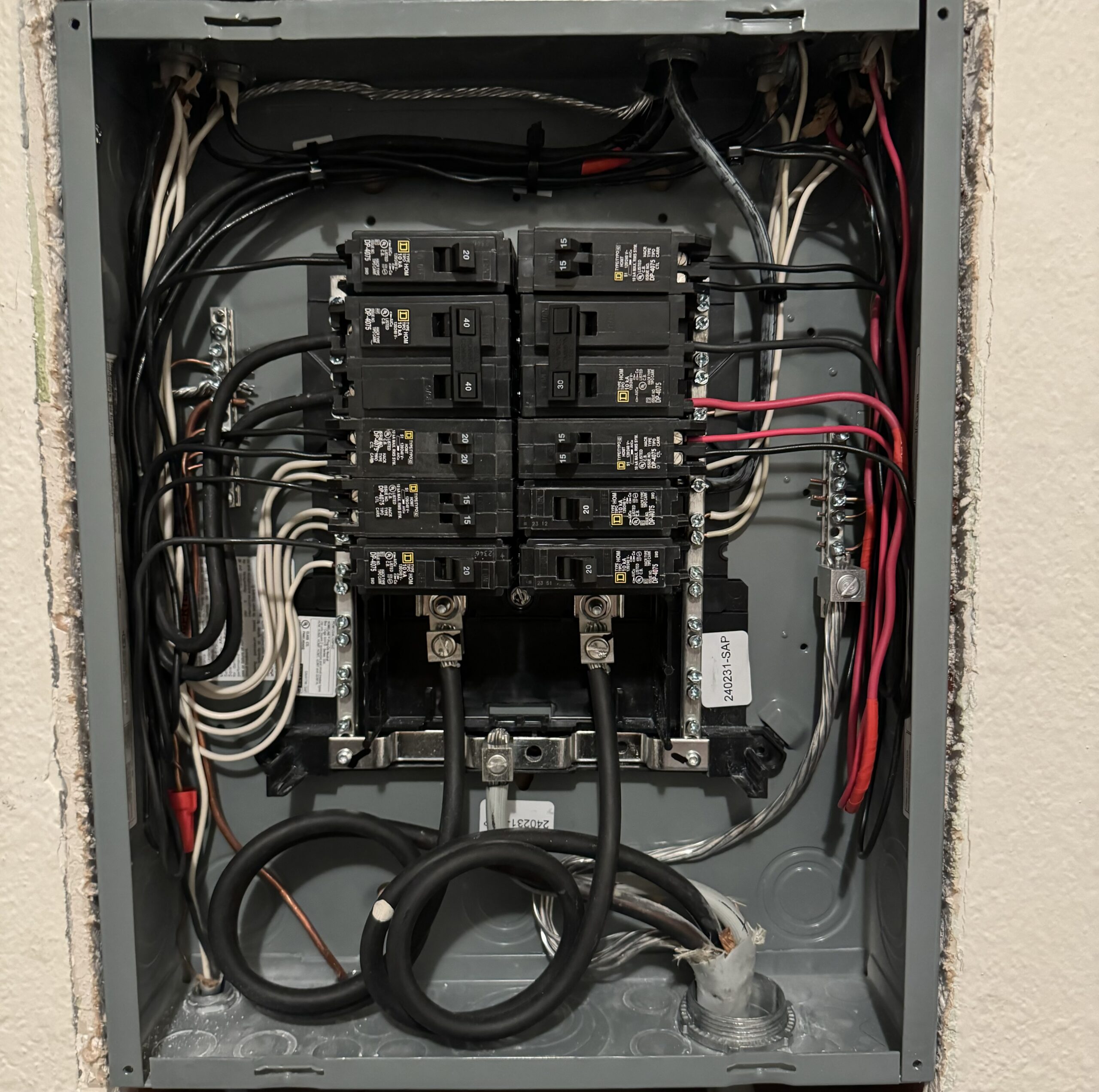 An electrical breaker panel with multiple circuit breakers, black and red wires, neutral wires on bus bars, and grounding wires, mounted on a wall.