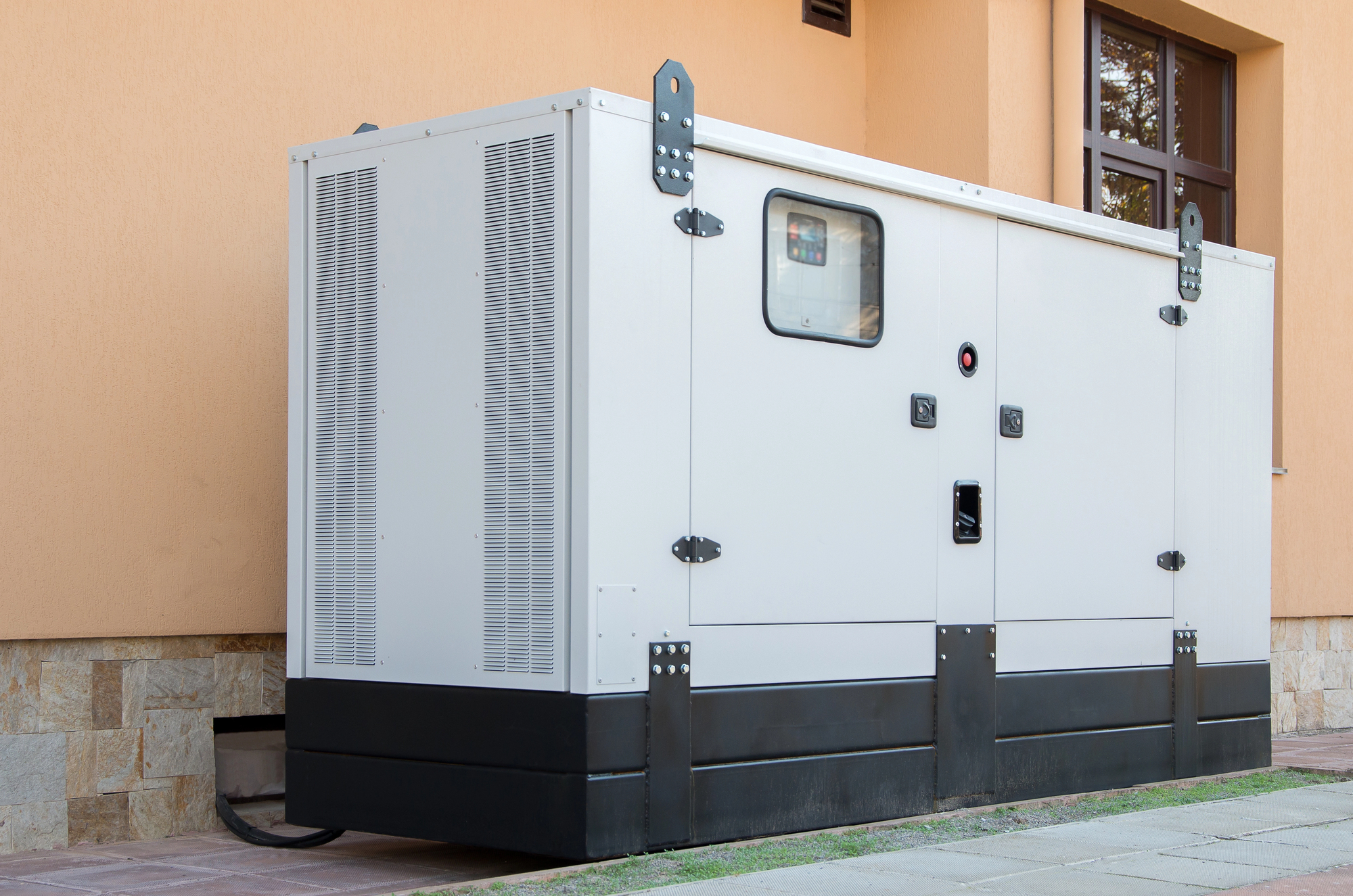 A large white standby generator is installed outside a building, positioned on a concrete slab near a beige wall.