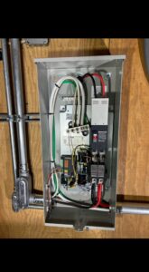 An open electrical panel mounted on a wood wall, showing various wires, circuit breakers, and electrical components inside.