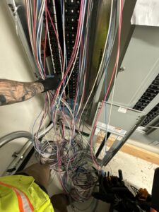 A person in a safety vest manages a bundle of colorful electrical wires in front of an open electrical panel.