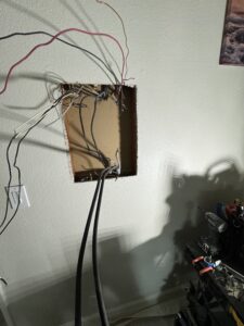 A wall with a rectangular hole exposing various electrical wires and cables, surrounded by tools and equipment in a dimly lit room.