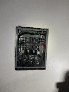 An open electrical panel is mounted on a white wall, showing circuit breakers and organized wiring inside.