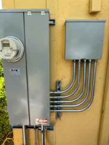 A gray electrical panel and meter are mounted on a yellow wall, with five metal conduit pipes running from the panel into a gray junction box.