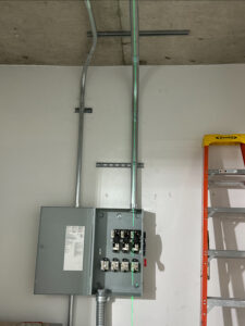 Gray electrical panel with open door mounted on a white wall, exposed conduit pipes above, and a partially visible orange ladder on the right.