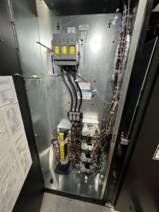 Open electrical control panel showing breakers, wiring, relays, and labeled components mounted on a metal backplate, with a schematic diagram attached to the door.
