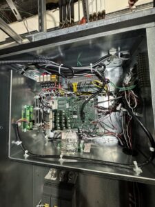 Open metal electrical panel showing a circuit board, various wires, connectors, and relays in an industrial setting.