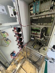 Open electrical control panel showing various circuits, wiring, relays, fuses, circuit boards, and electronic components mounted inside a metal enclosure.
