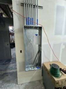An unfinished electrical panel with several conduits and wires installed, mounted on a drywall, with tools and materials nearby in a construction area.