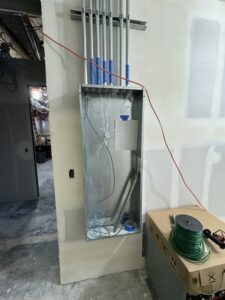 Electrical panel box with several conduit pipes and wires, some labeled with blue tape, installed on an unfinished wall in a construction area. Box contains wiring materials and tools.