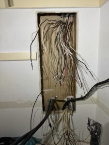 A wall panel with numerous exposed electrical wires of various colors hanging out, with no cover or organization.