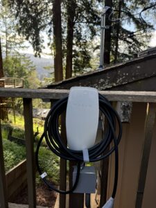 A Tesla wall connector for electric vehicle charging is mounted on a wooden fence with the cable neatly coiled. Trees and a house are visible in the background.