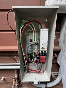 Open electrical panel mounted on a wall, showing various wires, circuit breakers, and electrical components inside the enclosure.