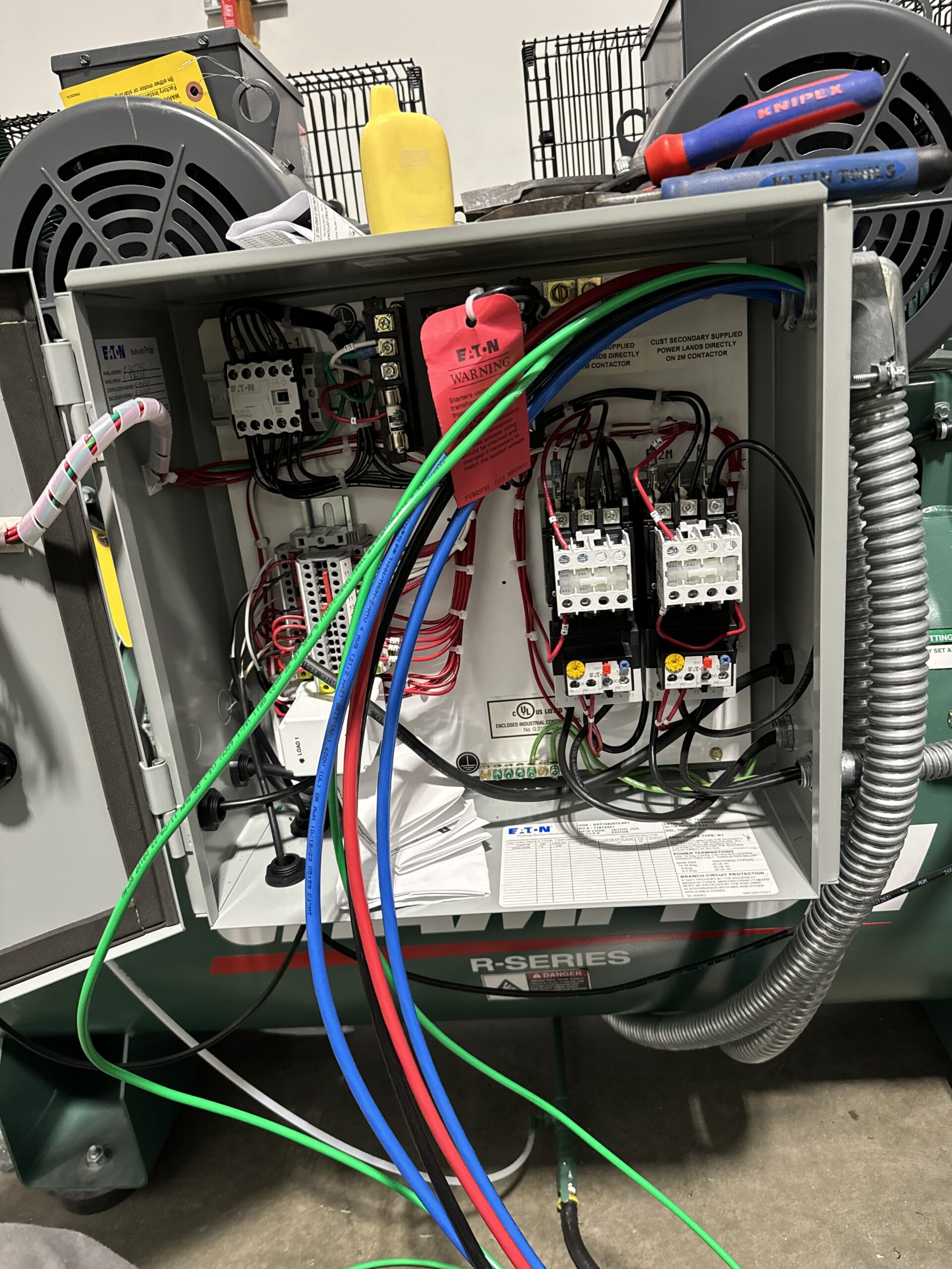 Open electrical control panel with exposed wiring, labeled tags, colorful cables connected, and electrical components visible inside a metal enclosure.